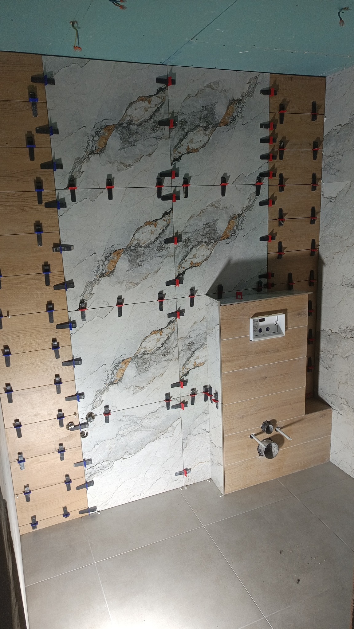 Ściany łazienki w trakcie układania płytek, widoczne krzyżyki dystansowe i fragment podłogi z jasnych płytek. Częściowo ułożona zabudowa WC.