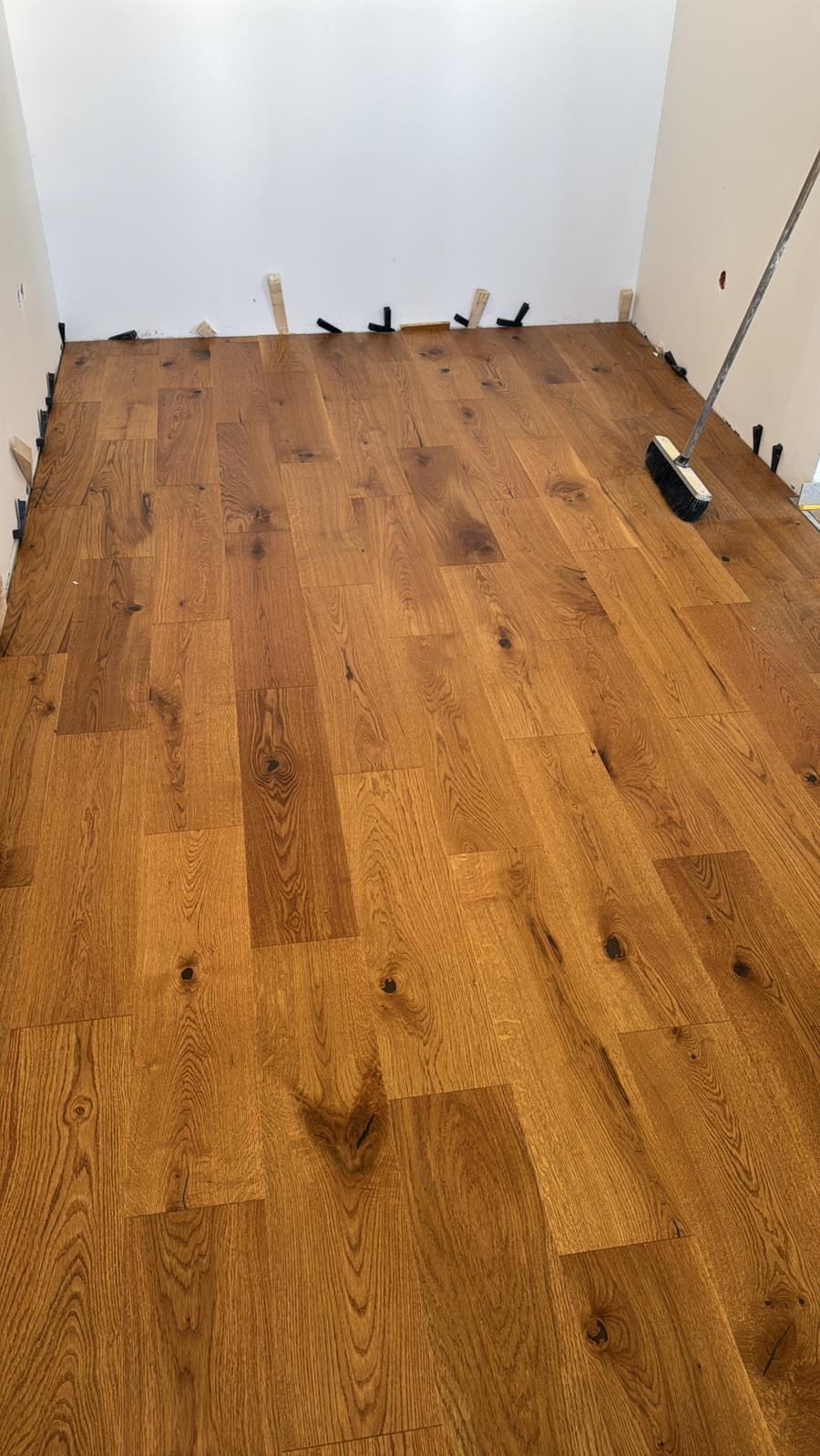 Ułożony parkiet dębowy w pokoju, widoczne kliny dystansowe przy ścianach i szczotka do zamiatania. Ciepłe odcienie drewna i naturalna faktura parkietu.