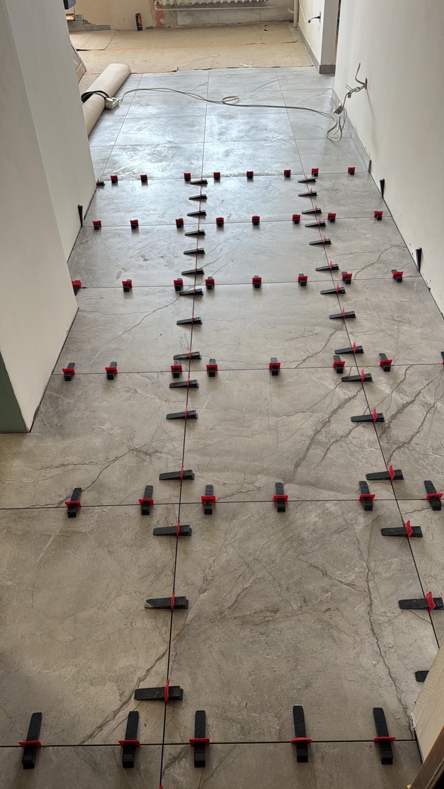 Szare płytki podłogowe z marmurkowym wzorem, wyrównane systemem poziomowania płytek z czarnymi klinami i czerwonymi klipsami, w trakcie układania na korytarzu.