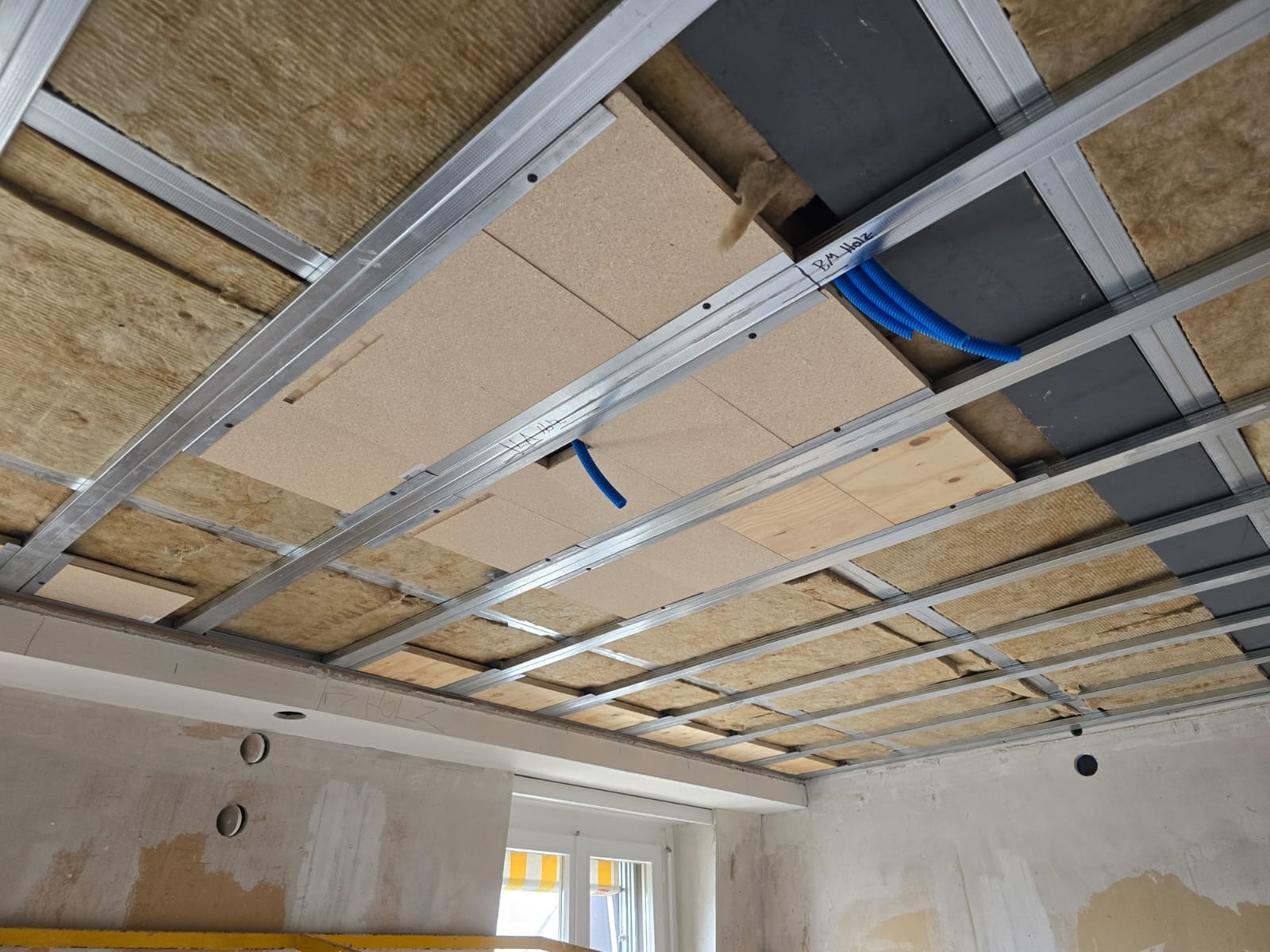 Sufit podwieszany w trakcie montażu: metalowa konstrukcja, izolacja z wełny mineralnej, fragmenty płyt. Widoczne instalacje elektryczne w peszlach.