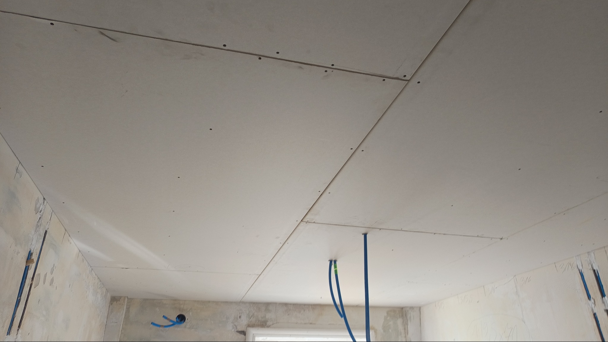 Zamontowane płyty karton-gips na suficie w trakcie remontu. Widoczne łączenia płyt i wystające rury instalacji. Surowe ściany w tle. Kąt patrzenia z dołu.