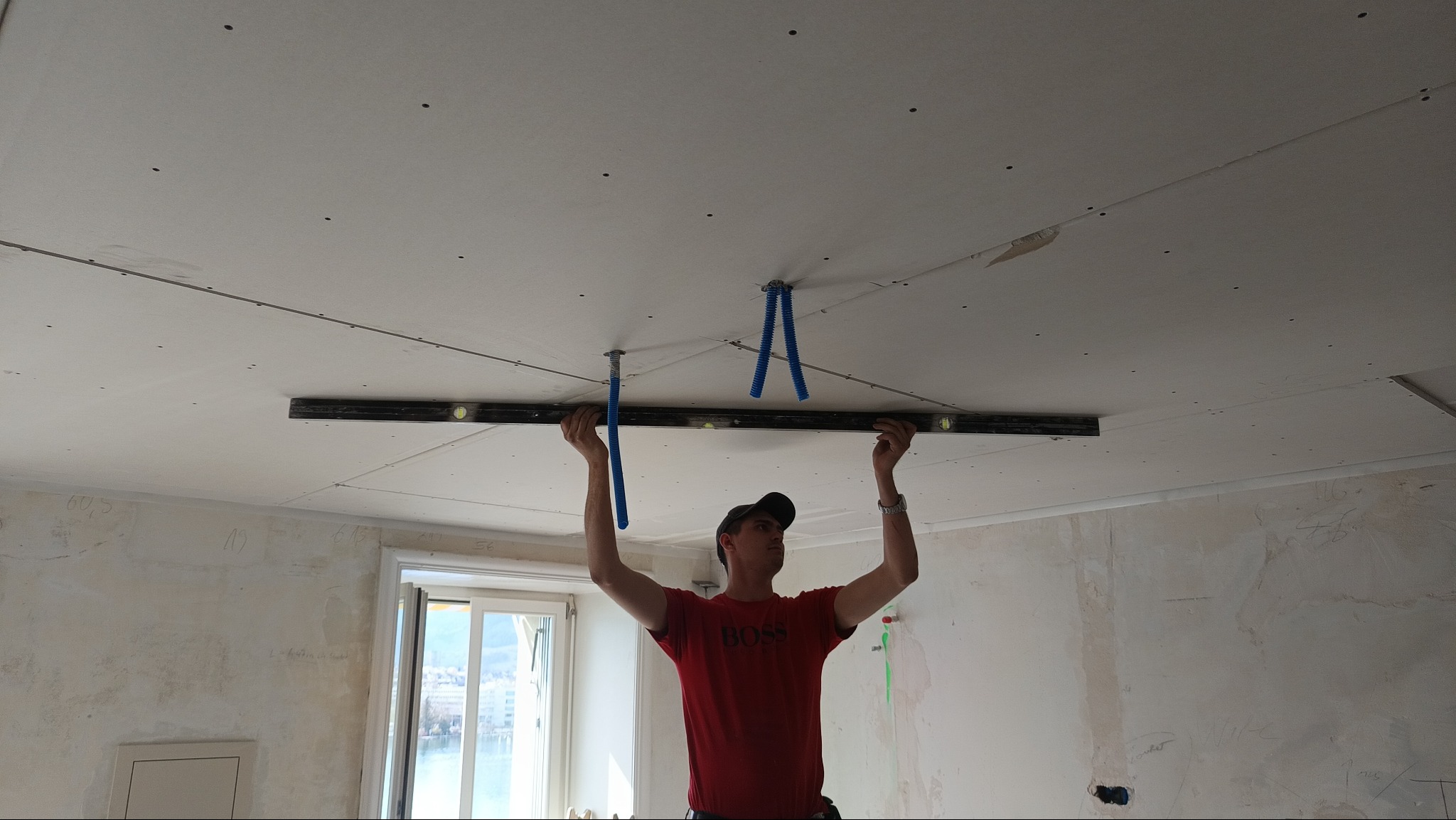 Pracownik z poziomicą przy montażu płyty gipsowej na suficie w pomieszczeniu z oknem. Widoczne elementy instalacji. Ściany w stanie surowym.