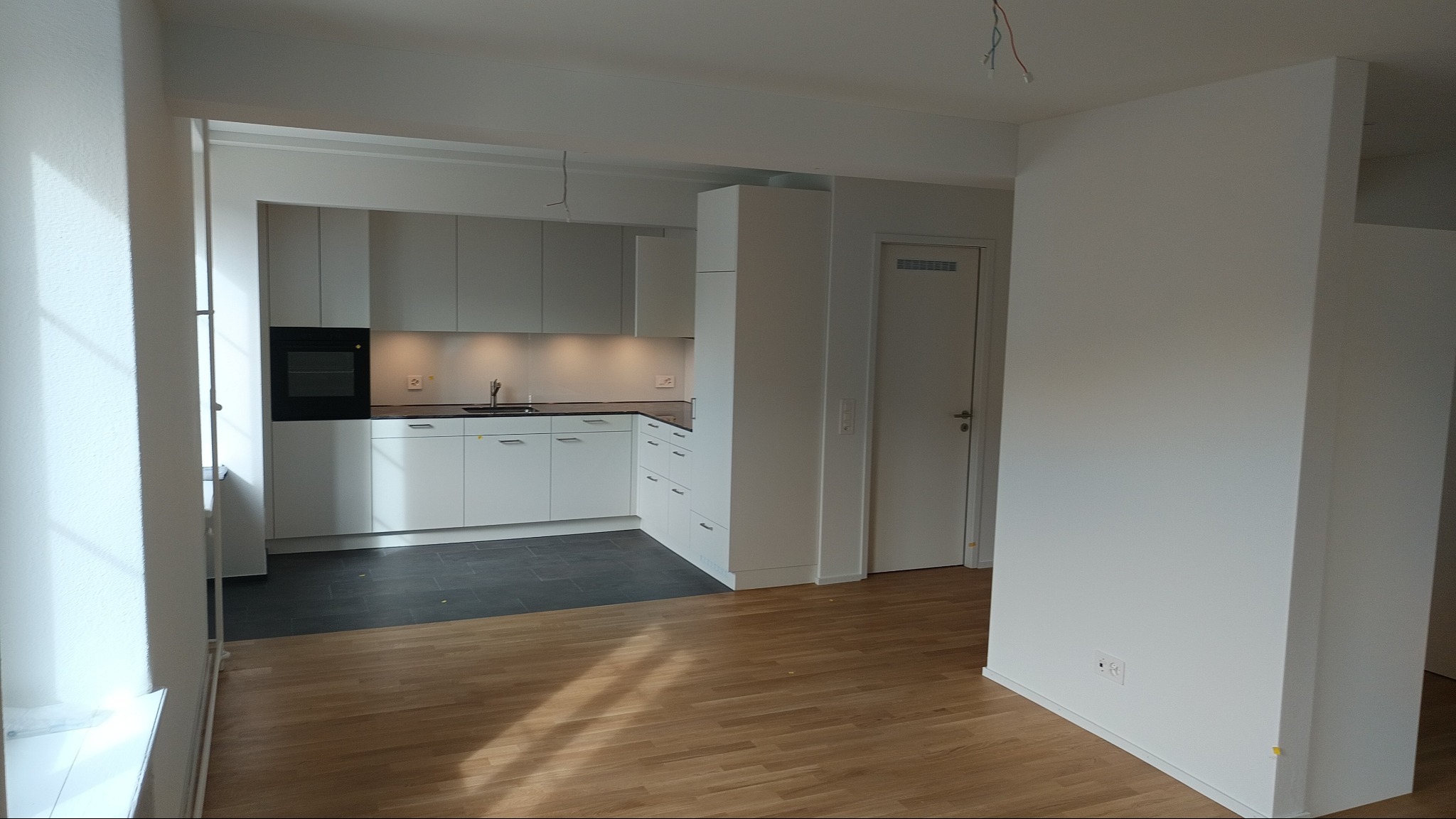 Jasna kuchnia z białymi szafkami i ciemnym blatem, widoczna z pokoju z parkietem. Minimalistyczne wnętrze z oknem po lewej stronie i elementami wykończenia.