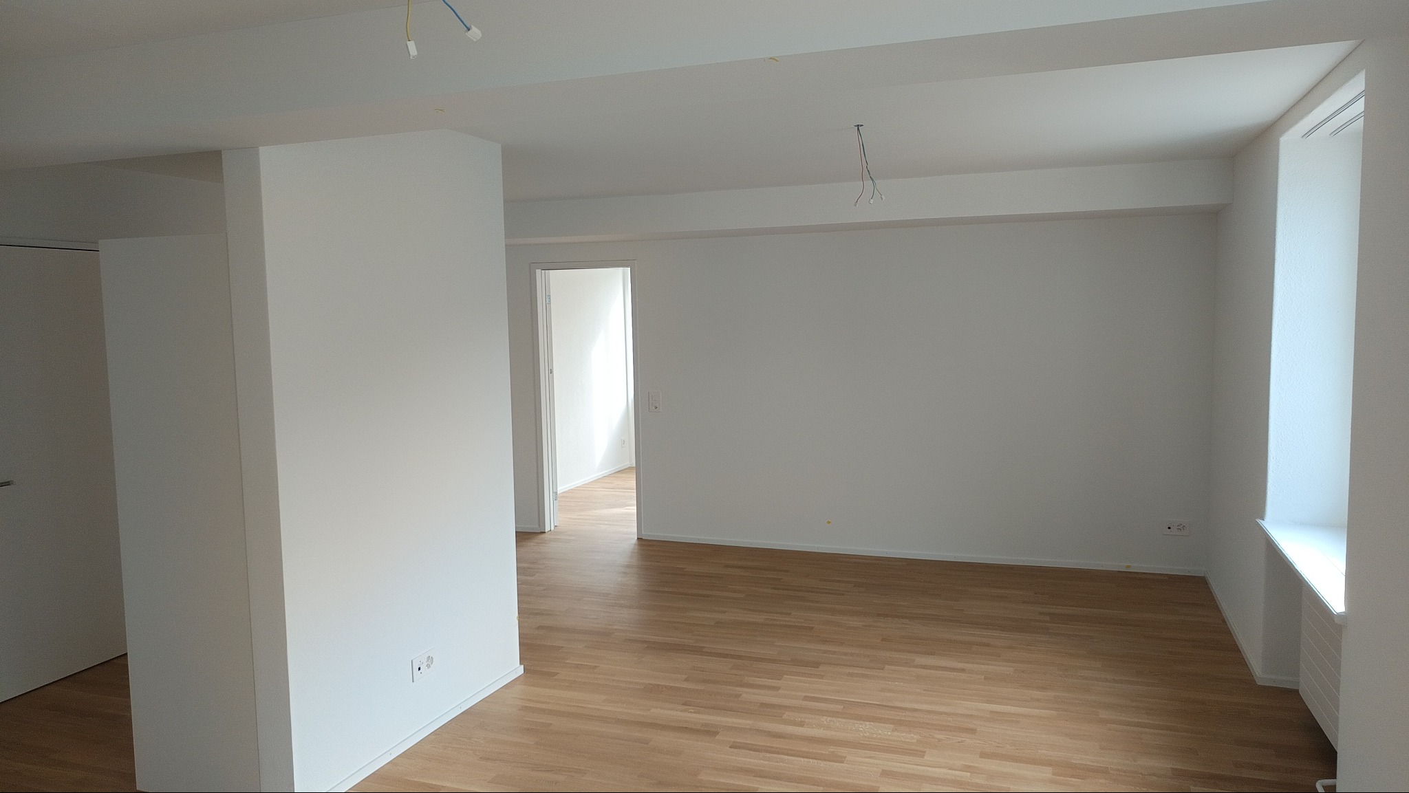 Jasne, przestronne wnętrze po remoncie, z parkietem i świeżo pomalowanymi ścianami na biało. Widoczne gniazdka elektryczne i surowe przewody oświetleniowe.