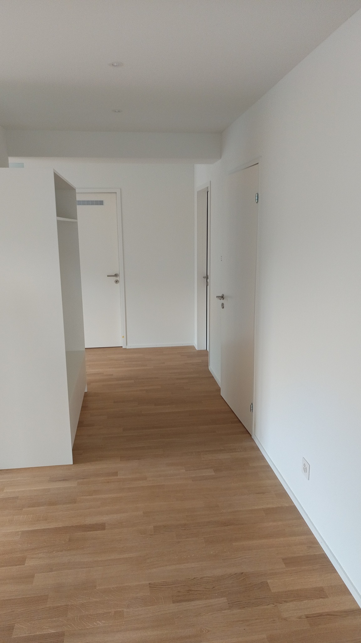 Jasne, odnowione wnętrze z drewnianą podłogą i białymi ścianami. Widoczne drzwi i fragment białej szafy. Minimalistyczny styl, świeżo po remoncie.