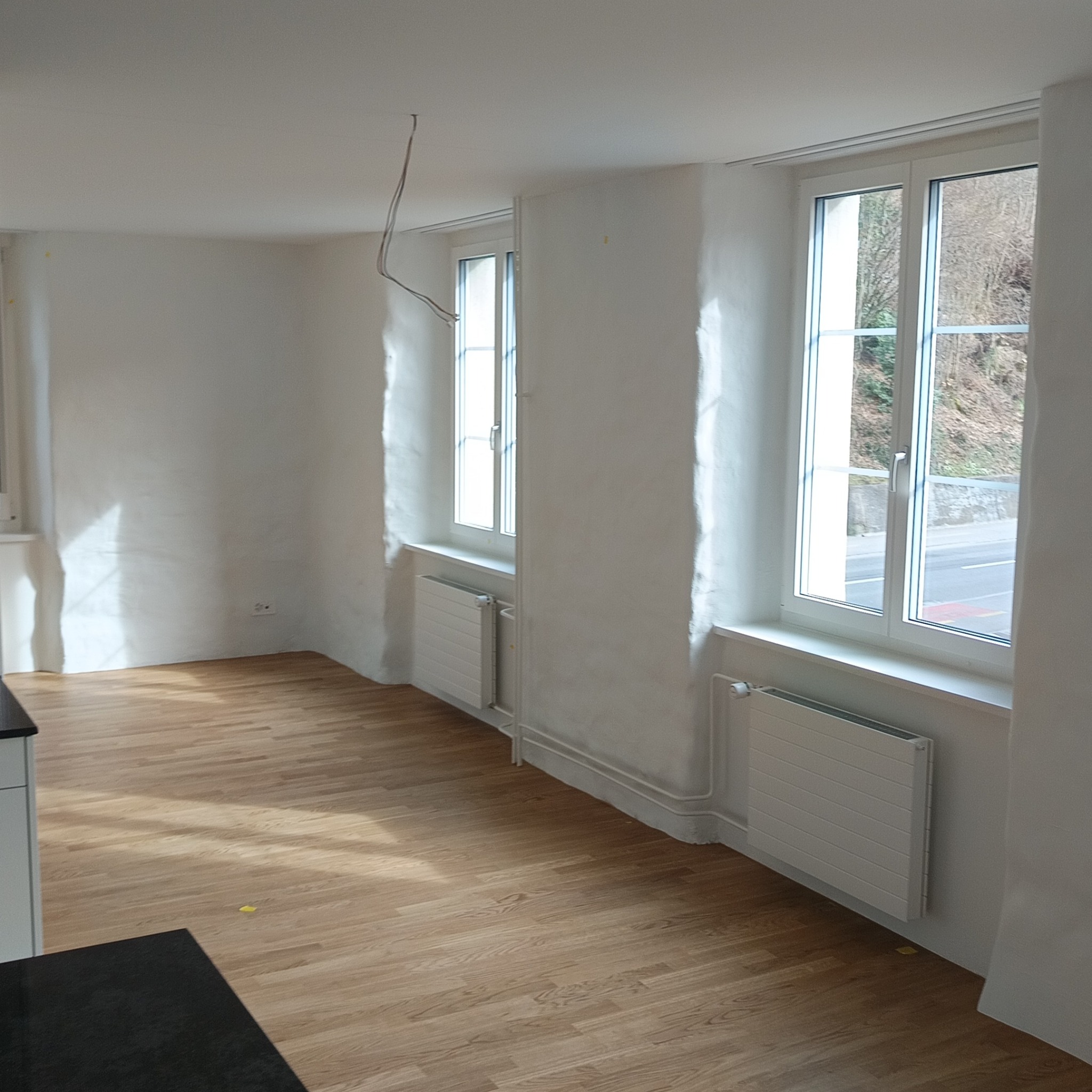 Jasne, odnowione wnętrze z drewnianą podłogą, białymi ścianami i grzejnikami pod oknami z widokiem na zieleń. Niedokończone przewody elektryczne zwisają z sufitu.