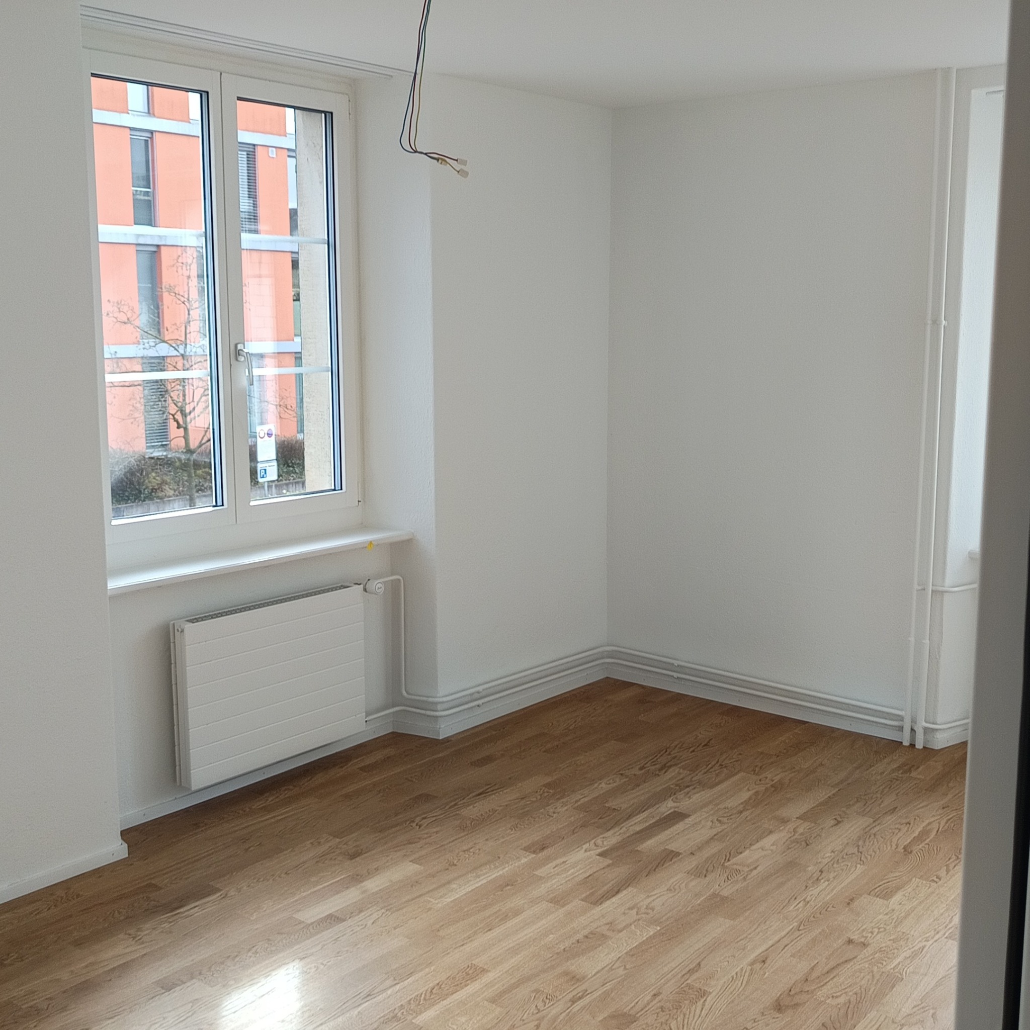Jasne, odświeżone wnętrze pokoju po remoncie: parkiet, białe ściany i grzejnik pod oknem z widokiem na budynek. Wiszące kable elektryczne w rogu.