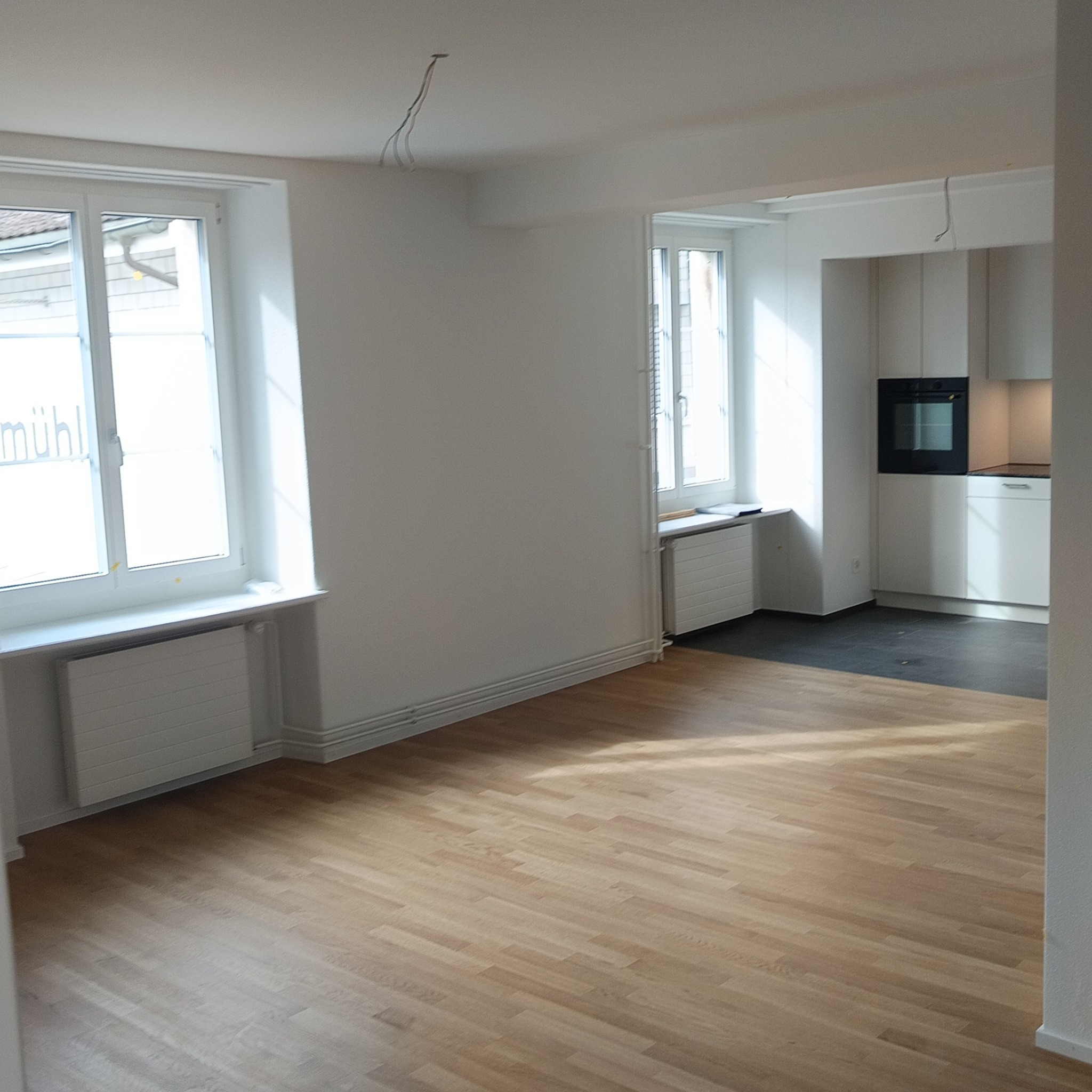 Jasne, przestronne wnętrze po remoncie z parkietem, białymi ścianami, grzejnikami pod oknami i widoczną kuchnią w tle. Wyprowadzone kable na suficie.