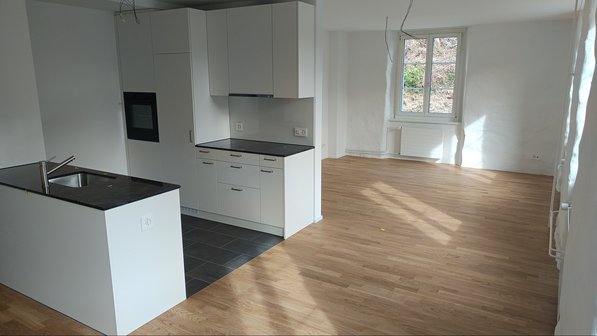 Jasna kuchnia z wyspą i drewnianą podłogą w nowym mieszkaniu. Minimalistyczny design, białe szafki i czarny blat. Widok na okno z zielenią w tle.