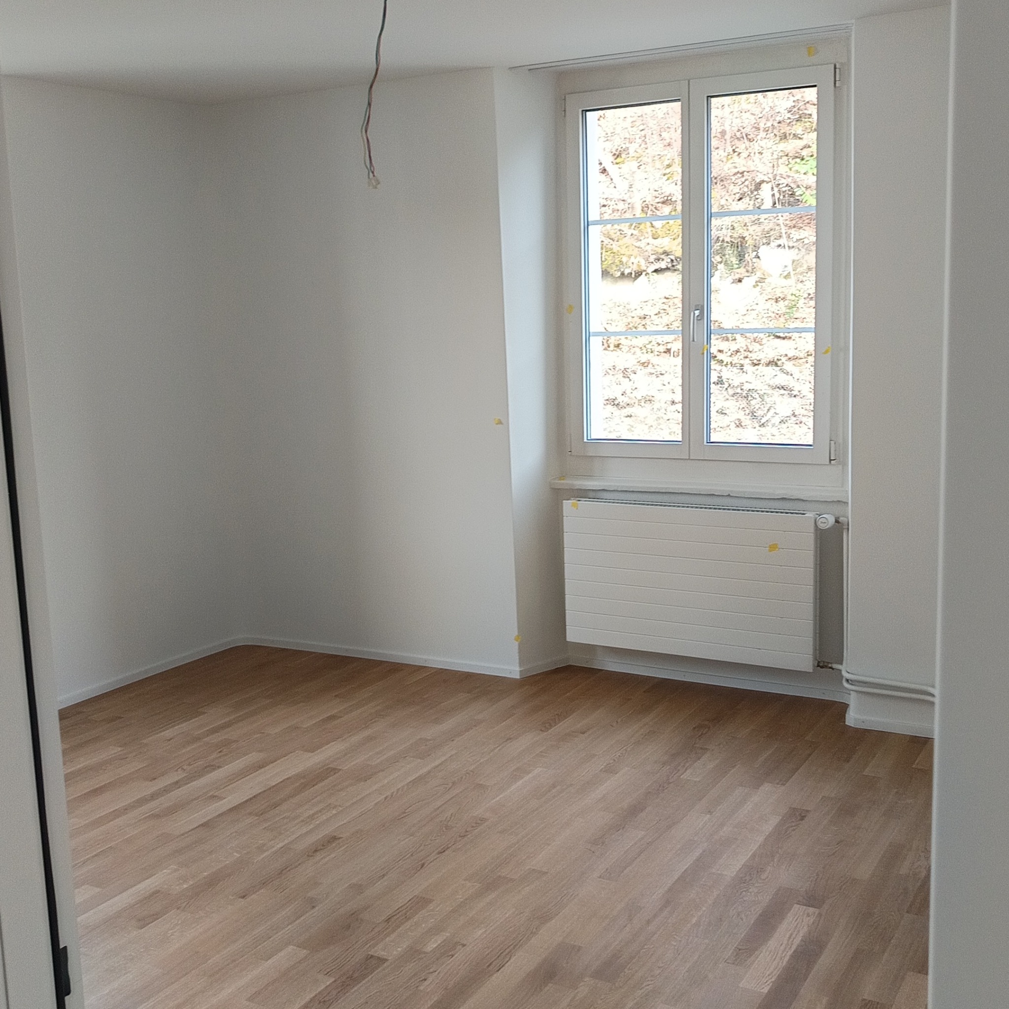 Jasne, puste wnętrze po remoncie: parkiet, białe ściany z widocznymi przewodami, okno z widokiem na drzewa. Żółte taśmy zabezpieczają elementy.