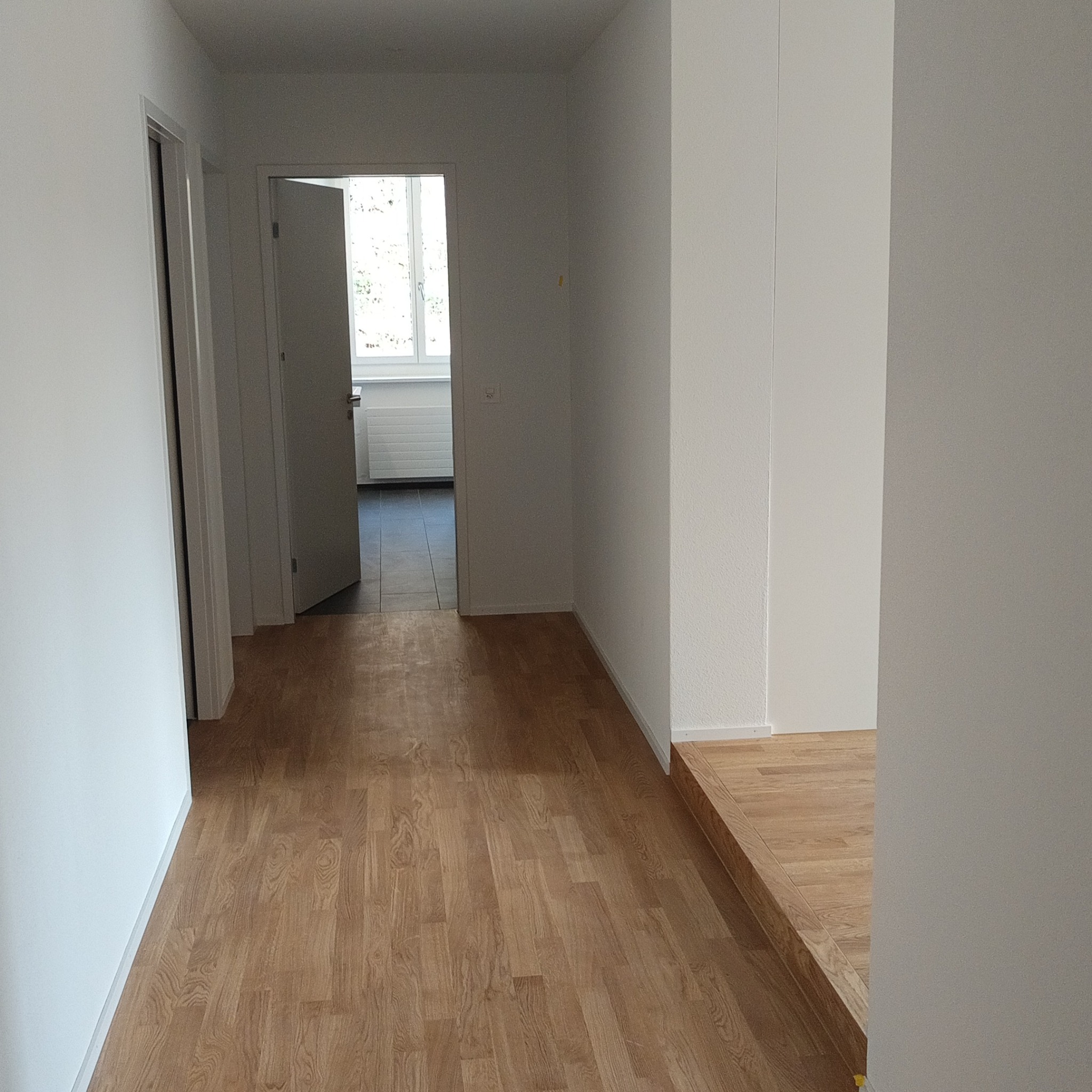 Jasny korytarz z parkietem i białymi ścianami. Widoczne otwarte drzwi do innego pomieszczenia z oknem. Minimalistyczny design, świeżo po remoncie.