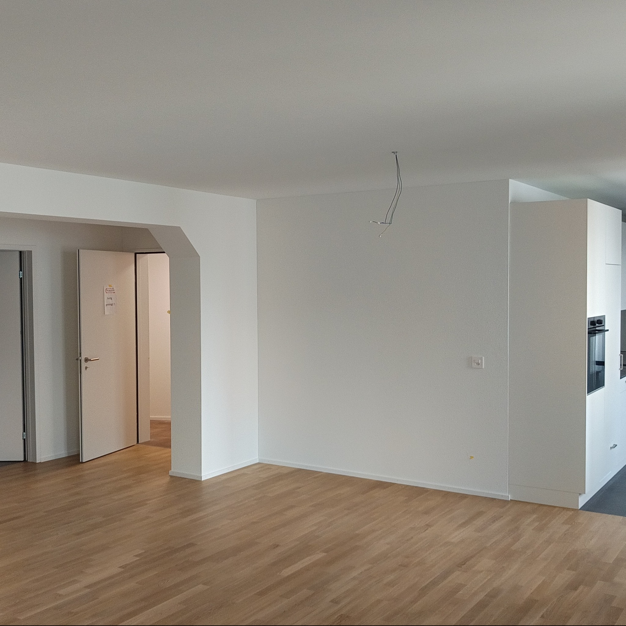 Jasne wnętrze po remoncie: parkiet, białe ściany, wnęka z kuchnią. Widoczne przewody oświetleniowe, minimalistyczny design, czyste linie i świeżość wykończenia.