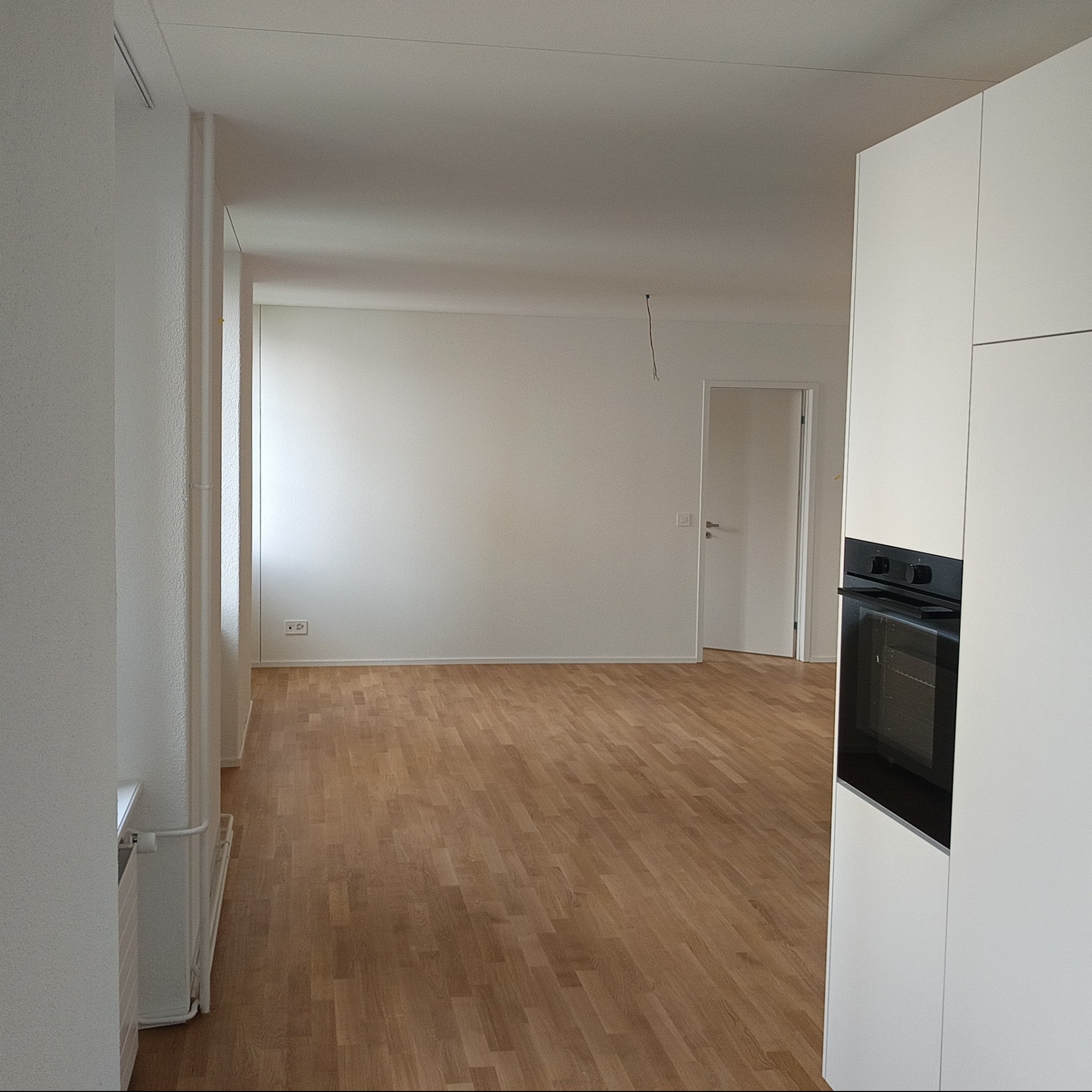 Jasne, przestronne wnętrze po remoncie z parkietem i białymi ścianami, widok na drzwi i fragment zabudowy kuchennej z piekarnikiem.