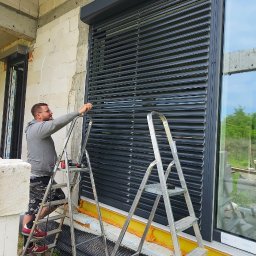 MIR TEAM MARIUSZ GRACZ, RAFAŁ TYMEK S.C. - Montaż żaluzji fasadowych na surowej elewacji budynku. Mężczyzna na drabinie reguluje lamele. Widoczne okno i narzędzia pracy.