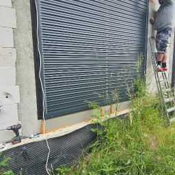 MIR TEAM MARIUSZ GRACZ, RAFAŁ TYMEK S.C. - Montaż szarej rolety zewnętrznej na tle surowej ściany budynku. Widoczna drabina, narzędzia i trawa. Instalator poprawia roletę. Kąt ujęcia z dołu.