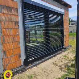 MIR TEAM MARIUSZ GRACZ, RAFAŁ TYMEK S.C. - Zamontowana, czarna żaluzja fasadowa z widoczną elewacją budynku w trakcie budowy. W tle widoczny budynek mieszkalny i błękitne niebo.