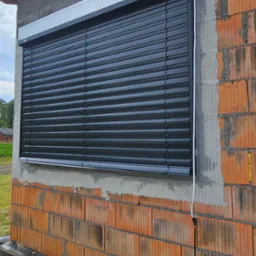 Ciemne rolety zewnętrzne zamontowane w oknie budynku w trakcie budowy, widoczne cegły i elementy instalacji elektrycznej na zewnątrz.