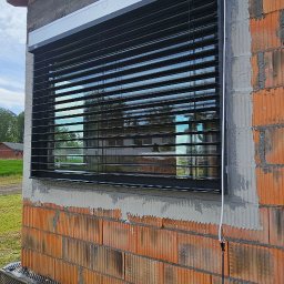 MIR TEAM MARIUSZ GRACZ, RAFAŁ TYMEK S.C. - Zamontowane żaluzje fasadowe w nowym budynku z cegły, widoczne odbicia w lamelach, kabel elektryczny zwisający z boku, żółty przedłużacz na ziemi.