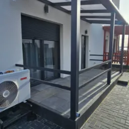 Nowoczesna pergola metalowa z zadaszeniem z poliwęglanu, szare słupki i balustrada. W tle okno z roletą zewnętrzną i klimatyzator na ścianie budynku.