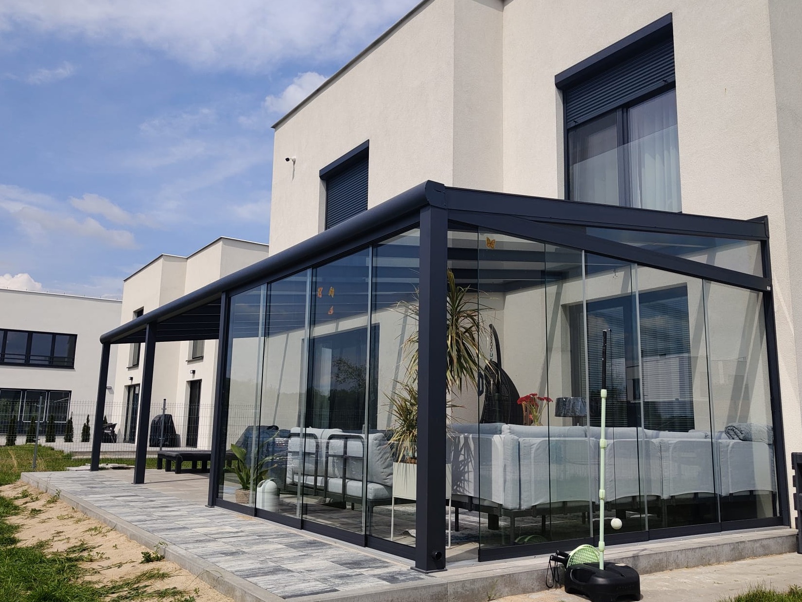 Nowoczesna, przeszklona pergola z grafitową ramą, przylegająca do jasnego budynku. Wewnątrz meble ogrodowe i rośliny. Widoczne rolety zewnętrzne na oknach budynku.