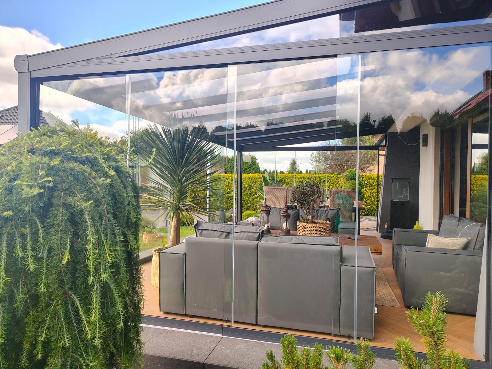 Nowoczesna, przeszklona pergola metalowa z przesuwnymi panelami szklanymi, widok z zewnątrz na komfortowe meble ogrodowe. W tle zieleń ogrodu.
