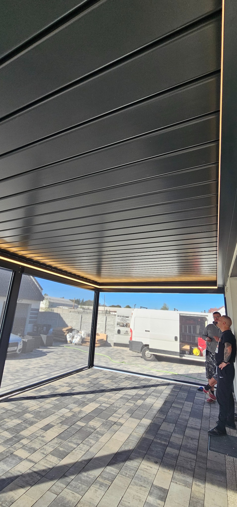 Nowoczesna pergola z metalu z oświetleniem LED, widok z wnętrza. Widoczne dwie osoby i samochód dostawczy na zewnątrz. Szara kostka brukowa na podłodze.