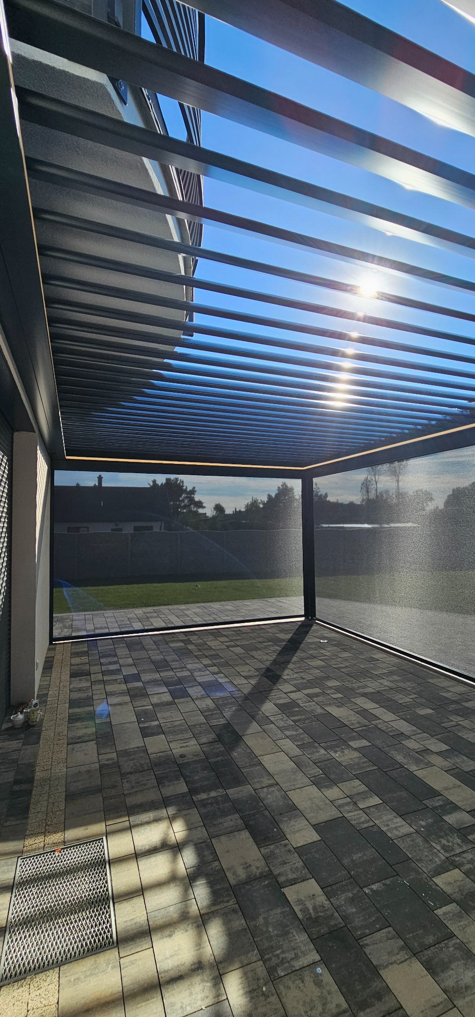 Nowoczesna, metalowa pergola z zasłoniętymi bokami, oświetleniem LED i widokiem na ogród w słoneczny dzień. Ciemna konstrukcja, jasne niebo, kostka brukowa na podłożu.