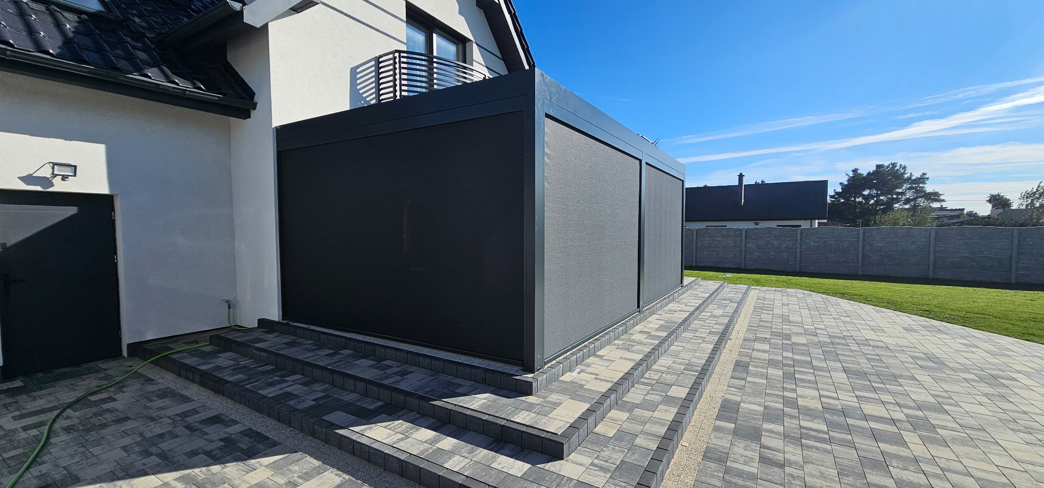 Nowoczesna pergola z ciemnymi roletami zewnętrznymi na tarasie z kostki brukowej, widok z boku w słoneczny dzień. Elementy domu i ogrodu w tle.