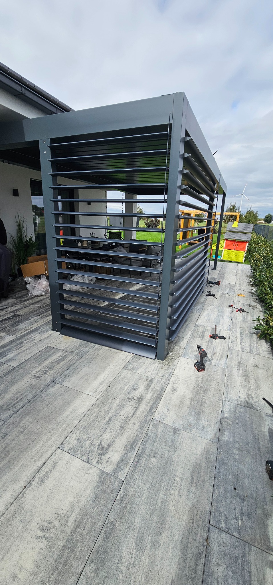 Szara, metalowa pergola lamelowa w trakcie montażu na tarasie z szarych płyt. Widoczne narzędzia i elementy konstrukcyjne. W tle dom i zielony ogród.