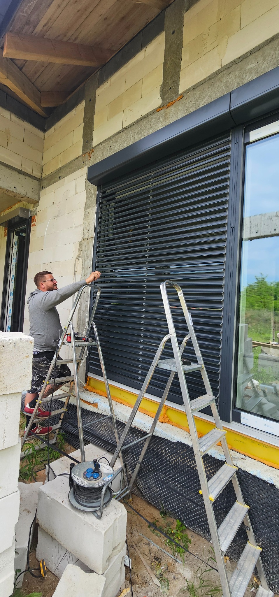 Montaż żaluzji fasadowych na surowej elewacji budynku. Mężczyzna na drabinie reguluje lamele. Widoczne okno i narzędzia pracy.