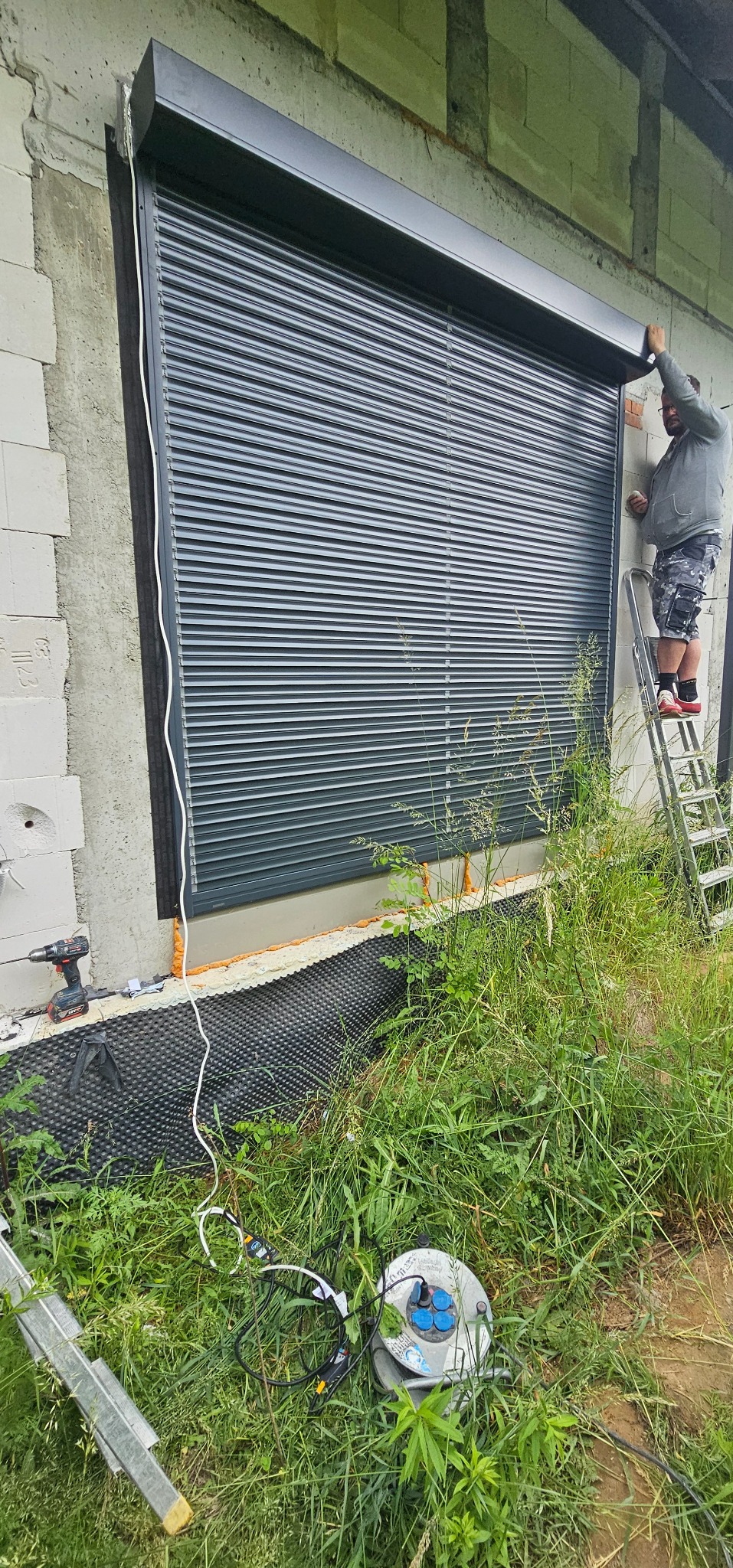 Montaż szarej rolety zewnętrznej na tle surowej ściany budynku. Widoczna drabina, narzędzia i trawa. Instalator poprawia roletę. Kąt ujęcia z dołu.
