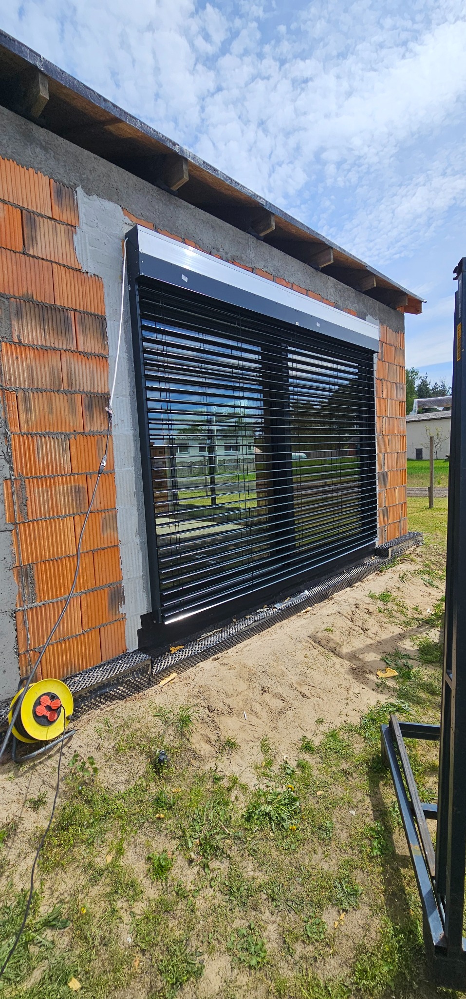 Zamontowana, czarna żaluzja fasadowa z widoczną elewacją budynku w trakcie budowy. W tle widoczny budynek mieszkalny i błękitne niebo.