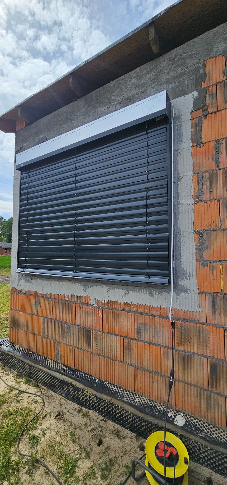 Ciemne rolety zewnętrzne zamontowane w oknie budynku w trakcie budowy, widoczne cegły i elementy instalacji elektrycznej na zewnątrz.