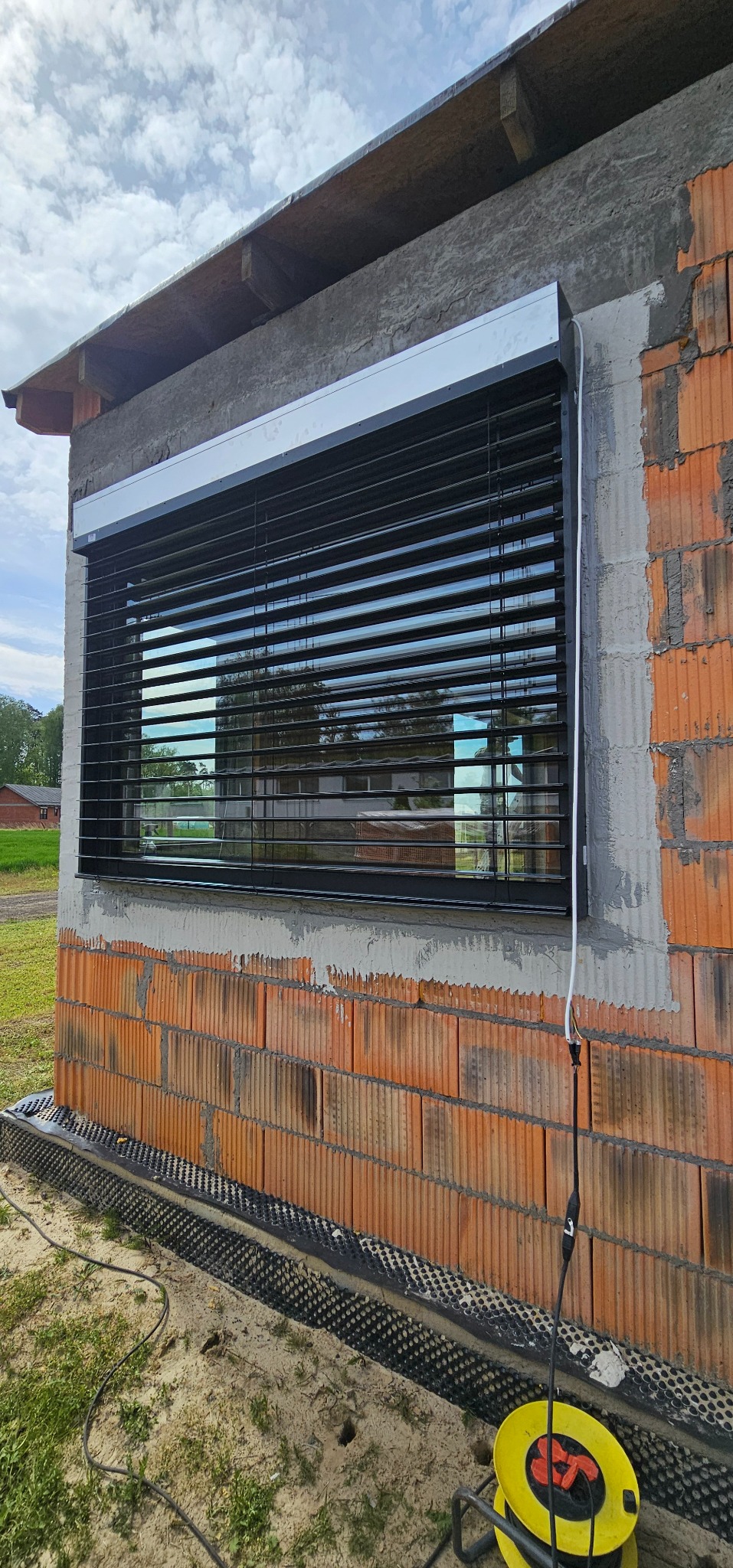 Zamontowane żaluzje fasadowe w nowym budynku z cegły, widoczne odbicia w lamelach, kabel elektryczny zwisający z boku, żółty przedłużacz na ziemi.