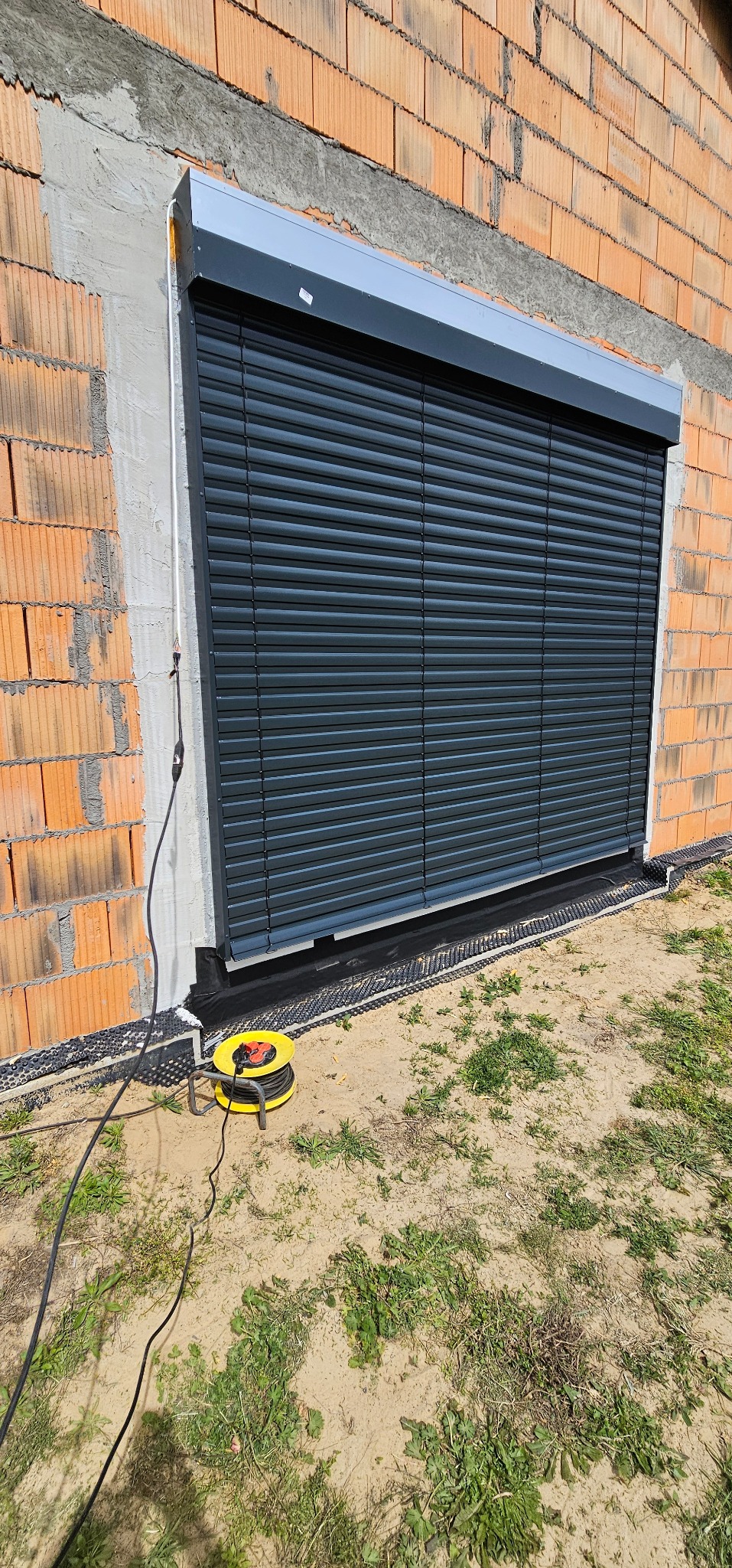Zamknięta, grafitowa roleta zewnętrzna na tle surowej ściany z czerwonej cegły i nieotynkowanych fragmentów. Widoczny zwinięty przedłużacz na ziemi.
