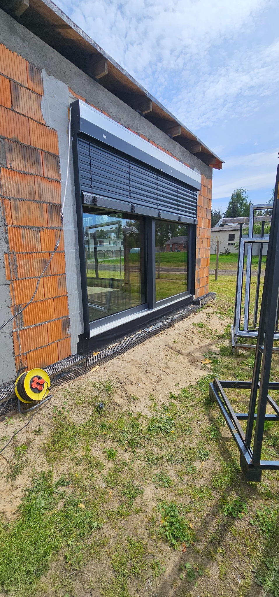 Nowe okno PCV z czarną roletą zewnętrzną na surowej ścianie budynku z cegły. Widoczne elementy montażowe i zwinięty przedłużacz. Częściowo odsłonięta elewacja.