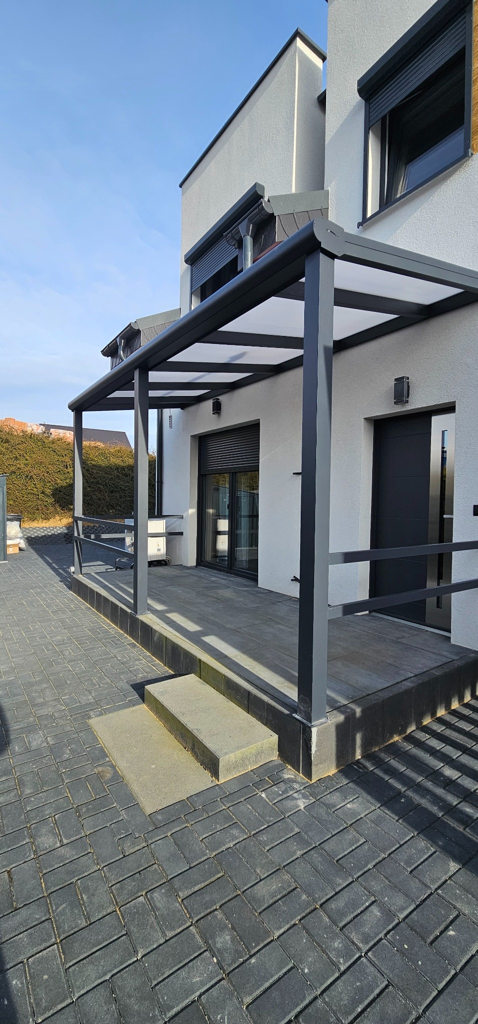 Nowoczesna, grafitowa pergola metalowa z poliwęglanowym dachem przy białym domu. Widoczne rolety zewnętrzne i kostka brukowa w jodełkę. Słoneczny dzień.
