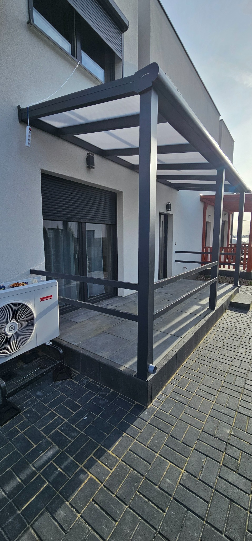 Nowoczesna pergola metalowa z zadaszeniem z poliwęglanu, szare słupki i balustrada. W tle okno z roletą zewnętrzną i klimatyzator na ścianie budynku.