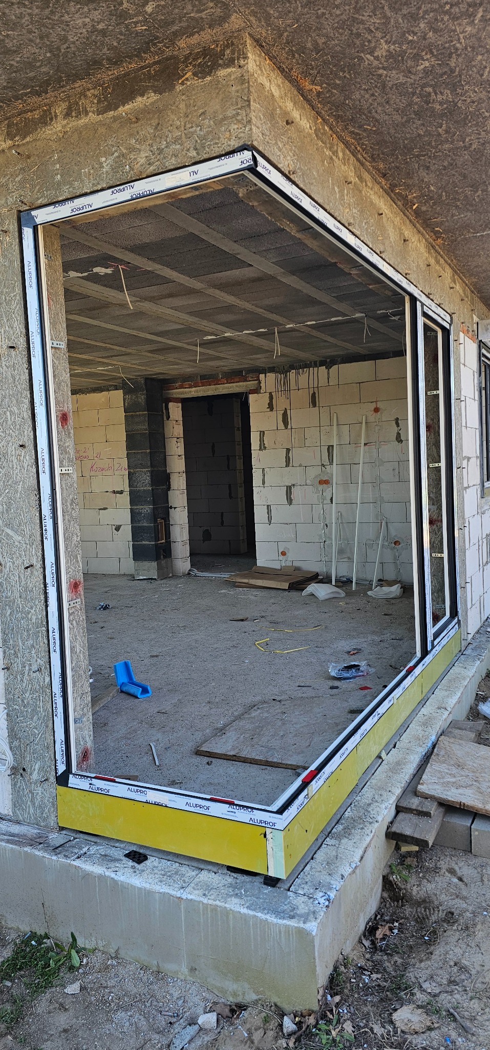 Wstawiony, ale nie wykończony, profil okienny Alupof w surowym otworze budynku w trakcie budowy. Widoczne surowe ściany z betonu komórkowego i strop.