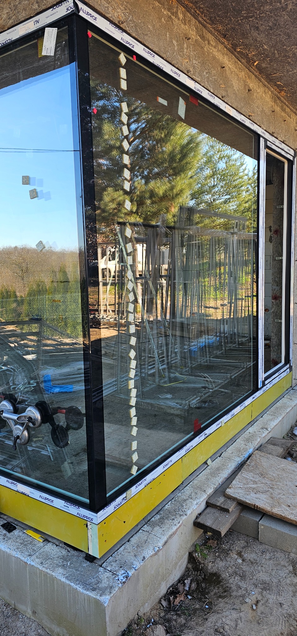 Narożne okno z czarną ramą i żółtą izolacją, widoczne narzędzia montażowe i rusztowania w tle. Na szybie przyklejone gąbki montażowe.
