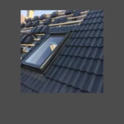 Częściowo pokryty dach z ciemnej dachówki z oknem dachowym; widoczne łaty i fragmenty niepokrytej połaci. Ujęcie z góry, w słoneczny dzień.