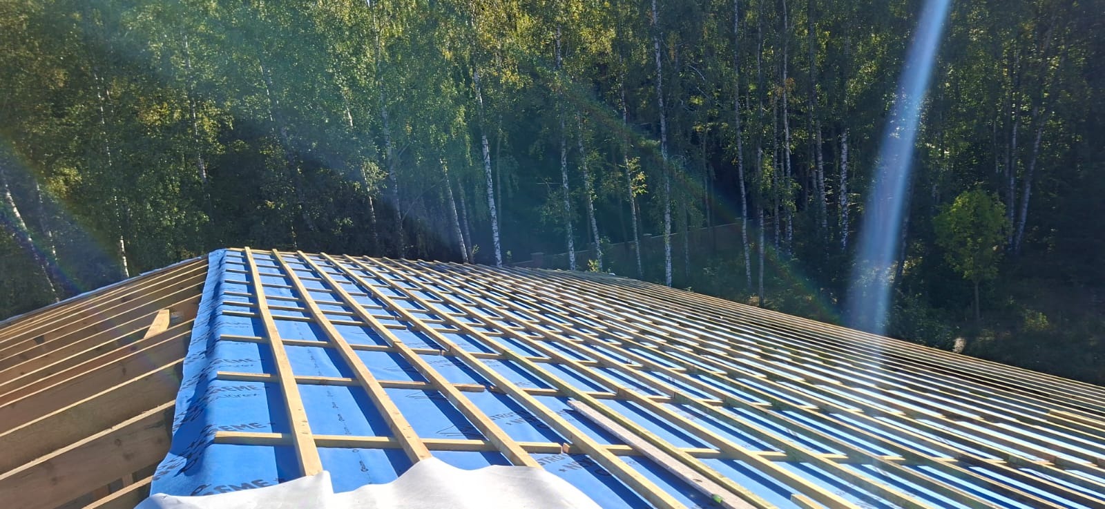 Wiązar dachowy z membraną w trakcie montażu na tle zieleni. Widoczne drewniane elementy konstrukcji i niebieska membrana dachowa. Promienie słoneczne w tle.