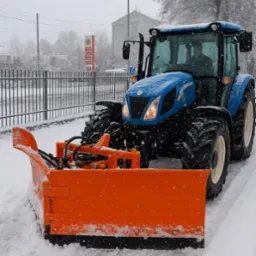 Niebieski traktor z pomarańczowym pługiem odśnieżającym na zaśnieżonej drodze; widoczne płatki śniegu, budynek w tle.