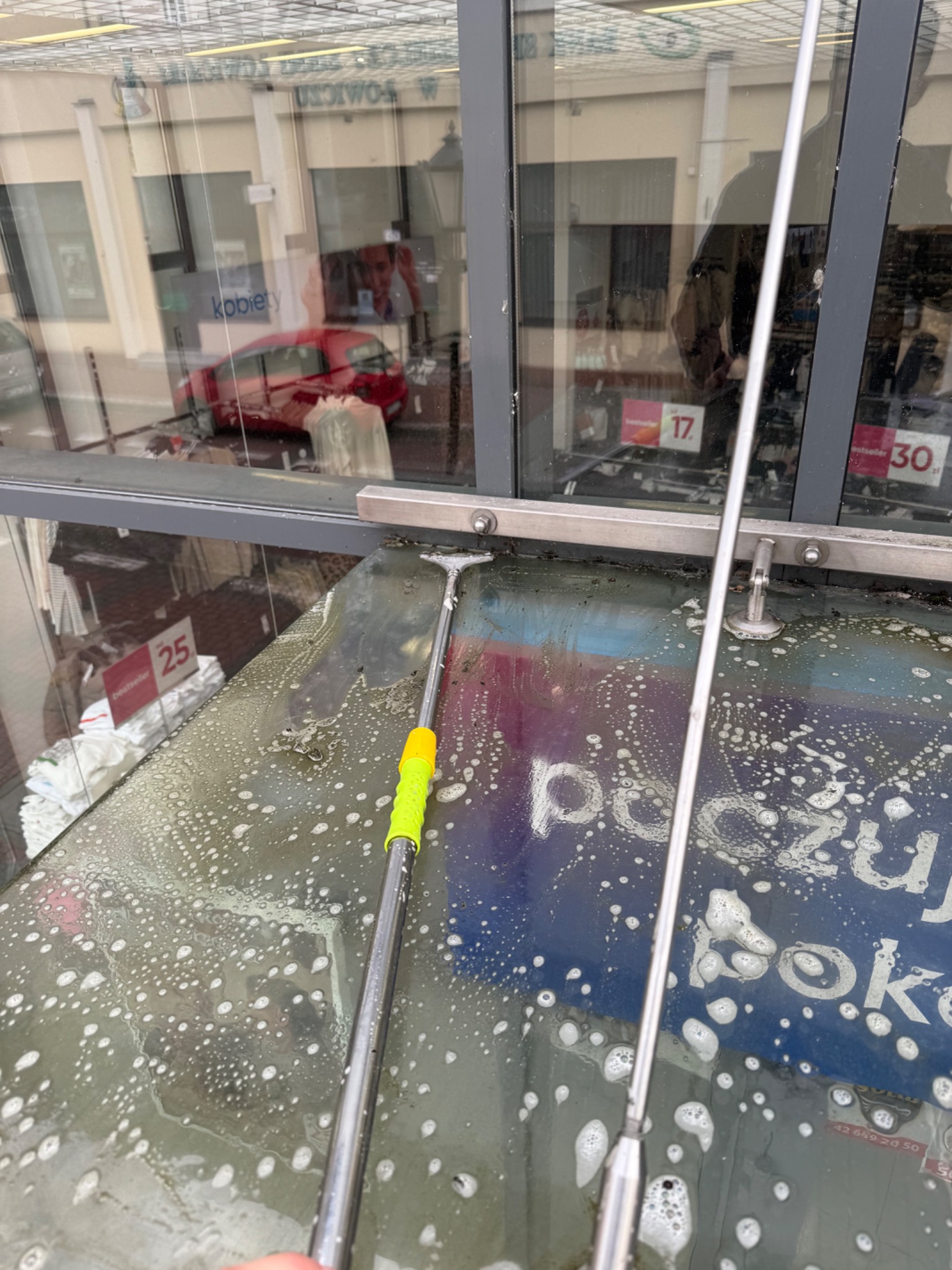 Mycie okna wystawowego z użyciem myjki teleskopowej; widoczne piana i refleksy otoczenia w szybie. Narzędzie z żółtym uchwytem. W tle widoczne towary w sklepie.
