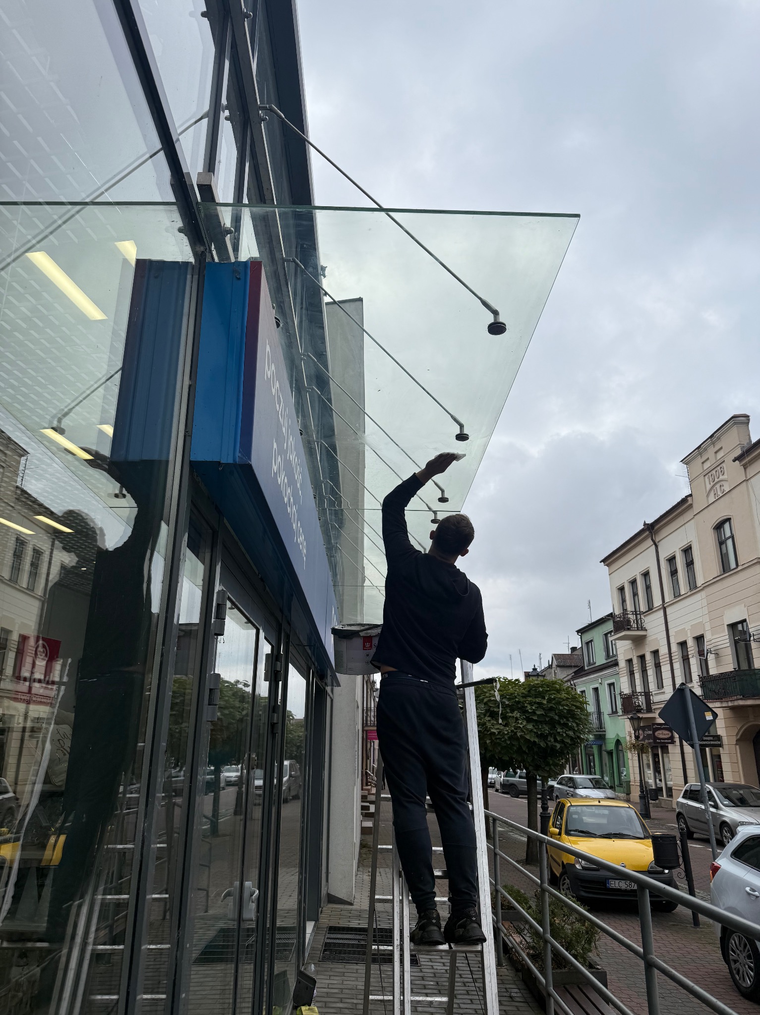 Mężczyzna na drabinie myje szklany daszek nad wejściem do budynku w miejskiej scenerii. Widoczna ulica i budynki w tle. Pochmurne niebo.