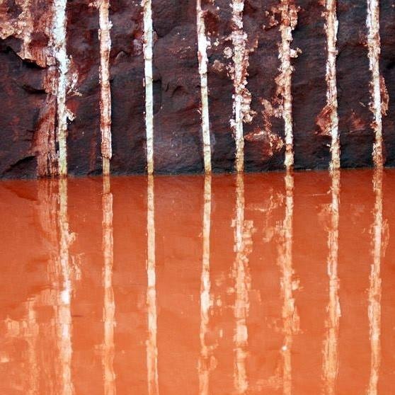 Abstrakcyjne ujęcie: pionowe białe linie na tle ciemnobrązowej powierzchni, odbijające się w pomarańczowej wodzie, tworząc efekt lustrzany i geometryczny wzór.