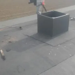 Montaż obróbki komina na dachu pokrytym papą, widoczna butla z gazem i narzędzia dekarskie.