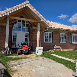 Markom Mariusz Komar - Budowa domu jednorodzinnego z cegły: białe okna z szprosami, czerwona dachówka, traktor-kosiarka i rower przed wejściem, błękitne niebo.