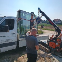 Markom Mariusz Komar - Montaż dużych okien z użyciem dźwigu przyssawkowego. Pracownicy w trakcie operacji na tle firmowego samochodu dostawczego i zabudowy jednorodzinnej.