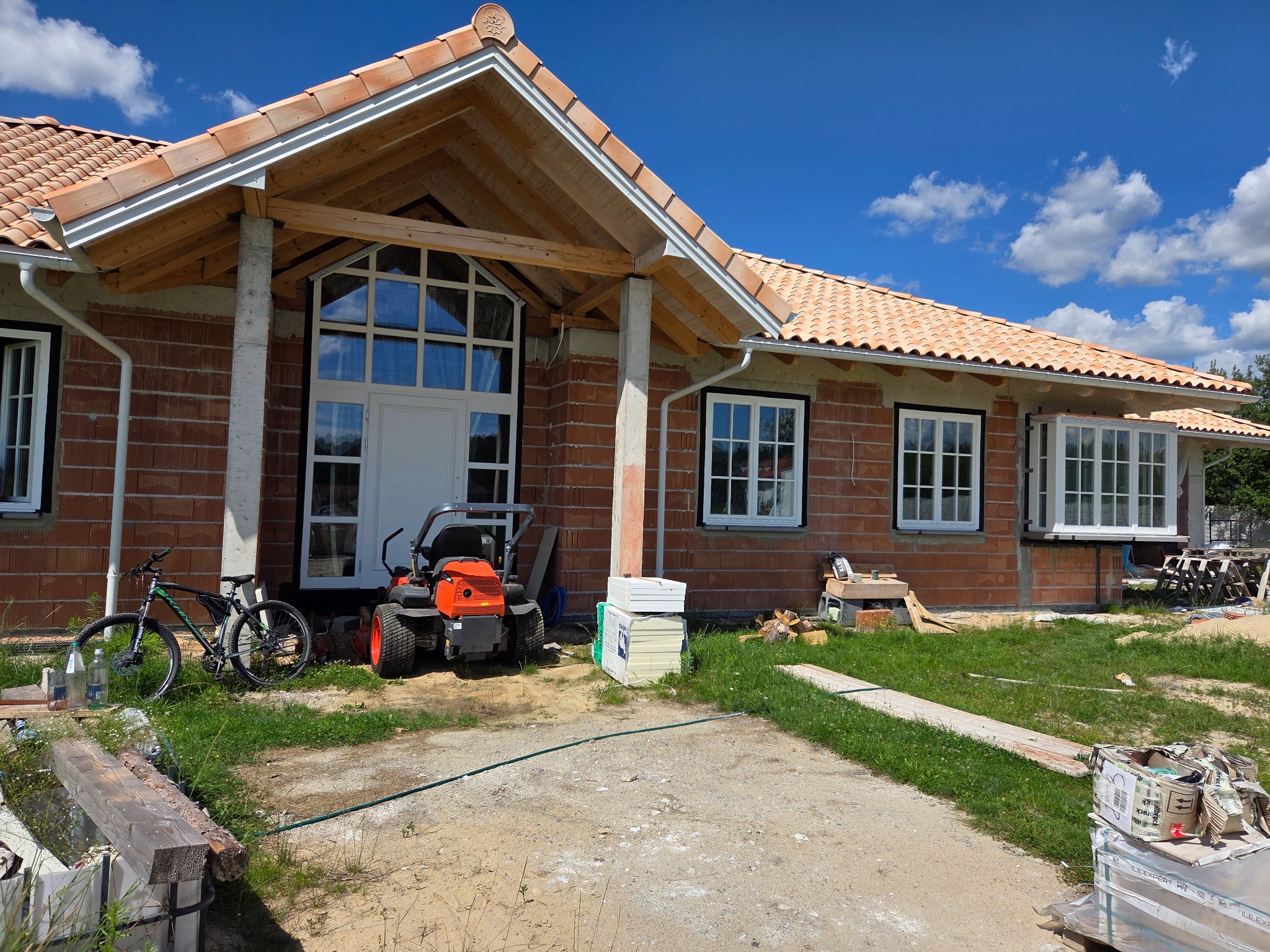 Budowa domu jednorodzinnego z cegły: białe okna z szprosami, czerwona dachówka, traktor-kosiarka i rower przed wejściem, błękitne niebo.